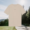 Sandbeiges Shirt mit dezentem kleinem Schriftzug, freigestellt vor sommerlicher Landschaft